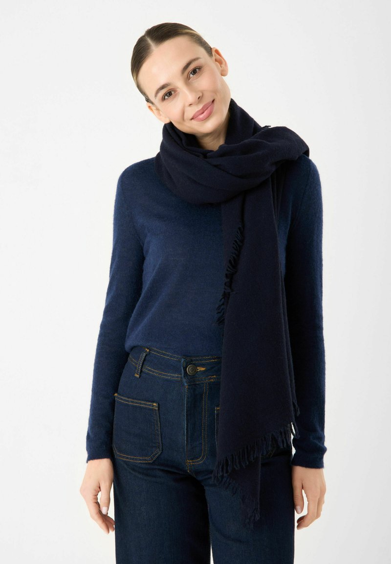 Sciarpa a maglia blu navy con bordi sfilacciati avvolta attorno al collo, abbinata a un top a maniche lunghe coordinato e jeans scuri con cuciture gialle.