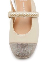 Zapato de piel beige con punta redondeada, adornado con una correa de perlas sintéticas blancas y una puntera plateada brillante.