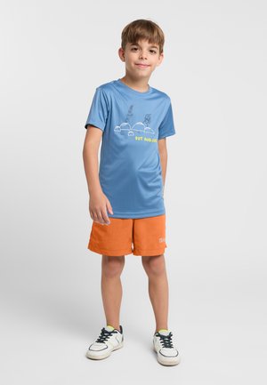 T-shirt bleue à manches courtes avec un design graphique et du texte jaune, associée à un short orange. Des baskets blanches avec des lacets noirs complètent la tenue.