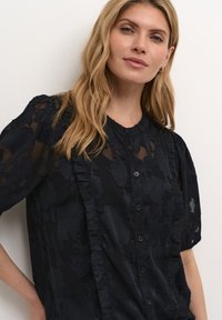 Blusa nera in chiffon con design a trama floreale, colletto arrotondato, parte frontale arricciata e maniche corte. Presenta bottoni davanti.