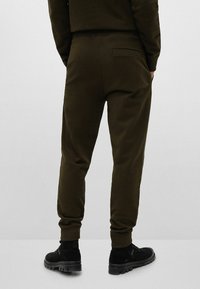 Olivegrüne Jogginghosen aus weichem Stoff, mit schmaler Passform, Bündchen an den Säumen und einer einzigen Gesäßtasche. Getragen mit schwarzen Schuhen.