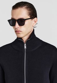 Marineblauer, gerippter Reißverschluss-Pullover mit hohem Kragen, silberner Reißverschluss, getragen mit dunkler Sonnenbrille, zeigt ein schlankes und modernes Design.