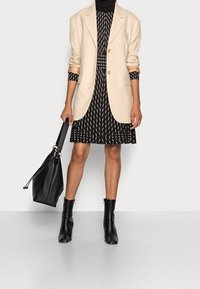 Frau in schwarzem gemustertem Kleid mit beigem Blazer, schwarzen Stiefeletten und schwarzer Handtasche an der Seite.