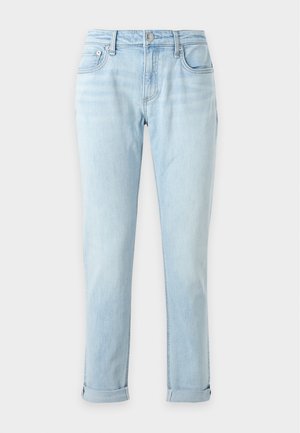 Jeune femme debout, une main dans la poche, portant une chemise jaune nouée sur un top rayé, un jean bleu clair roulé aux chevilles et des ballerines noires.