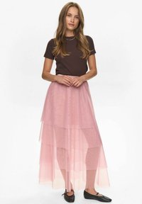 Mujer de pie con las manos juntas, vistiendo una blusa marrón de manga corta, una falda de tul rosa en capas y zapatos planos negros, contra un fondo blanco.
