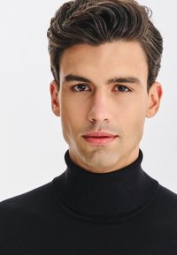 Młody mężczyzna z ciemnymi falującymi włosami, brązowymi oczami, ubrany w czarny golf, patrzący bezpośrednio w obiektyw na tle jednolitego białego tła.