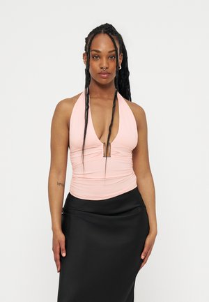 Donna con lunghe trecce che indossa un top rosa chiaro allacciato al collo con scollo a V profondo e gonna nera, in piedi davanti a uno sfondo bianco semplice.