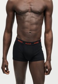 Buste masculin musclé à la peau foncée portant un boxer noir avec le logo rouge « HUGO » sur la taille, sur un fond uni.