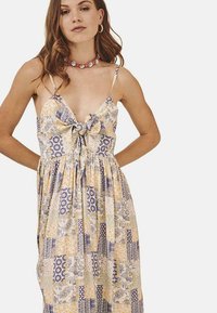 Vestido con estampado floral, con un lazo en la parte delantera, tirantes finos, tela ligera y colores amarillo, azul y blanco en un diseño de patchwork.