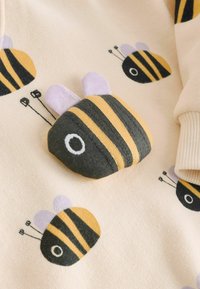 Mehak čebelji aplikacija z rdečimi, rumenimi in vijoličnimi črtami na bež tkanini z vzorcem čebel blizu rebraste manšete.