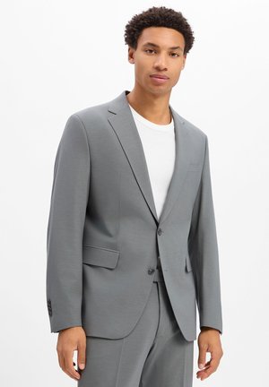 Man staat, draagt een lichtgrijze colbert en bijpassende broek met een wit t-shirt met ronde hals tegen een effen witte achtergrond.