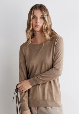 Kvinde med bølget blondt hår iført en lysebrun langærmet sweater, der holder brune læderstropper mod en ensfarvet hvid baggrund.