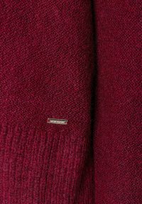 Burgunderfarbener Strickpullover aus strukturiertem Stoff, mit Rippenabschluss und einem kleinen metallischen Markenetikett an der Seite angenäht.