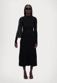 Musta ribbed midi mekko, jossa on pitkät hihat ja korkea kaulus, yhdistettynä mustiin nilkkureihin ja olkalaukkuun.