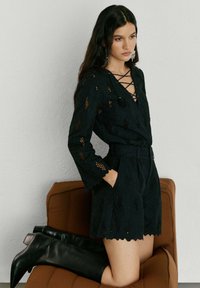 Sieviete melnā mežģīņu kombinzonā ar garām piedurknēm un krusteniskām kakla detaļām, atbalstoties pret brūnu krēslu, valkājot melnas ceļgalu augstus zābakus.