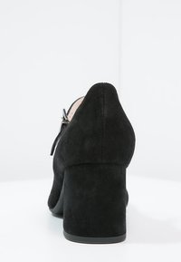 Chaussure à talon haut en suède noir avec un talon épais. L'arrière présente une texture lisse et une courbe subtile en haut. Pas de matériel visible.