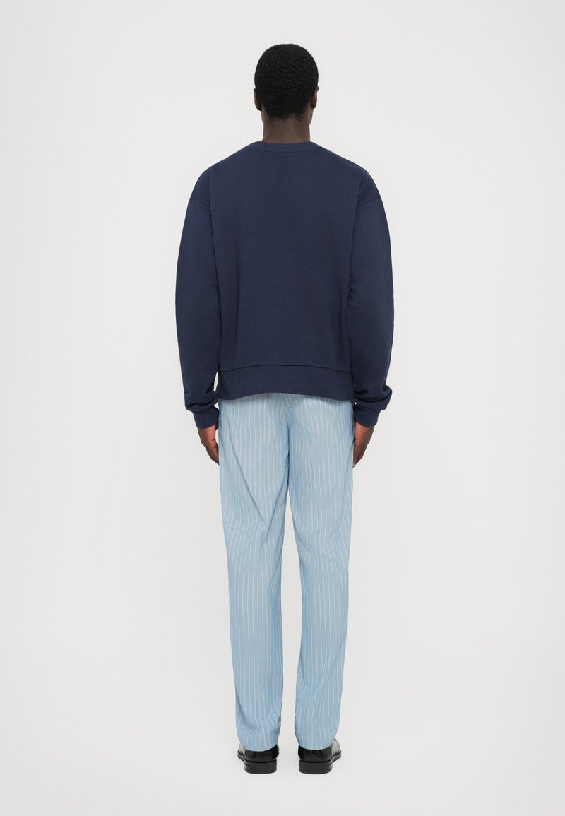 Navy crewneck sweatshirt with long sleeves paired with light blue pinstriped pants and black shoes, viewed from the back.