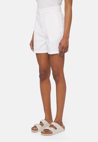 Pantaloni corti bianchi su misura con vita alta, fronte piatto e tasche laterali, abbinati a sandali grigio chiaro con due fibbie.