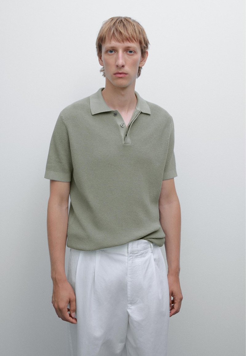 Polo shirt di colore verde oliva con texture, colletto e due bottoni, abbinato a pantaloni bianchi a gamba larga. Design semplice e moderno.
