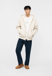 Νεαρός άνδρας που φοράει μπεζ ζακέτα με κουκούλα και φερμουάρ, λευκό μπλουζάκι Hollister, σκούρο μπλε τζιν και καφέ σουέντ μπότες μέχρι τον αστράγαλο, στέκεται με τα χέρια στις τσέπες.