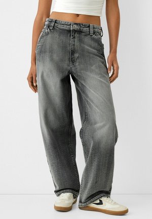 Personne portant un jean gris délavé à jambes larges, un crop top blanc et des baskets blanches avec des détails noirs, debout devant un fond uni.