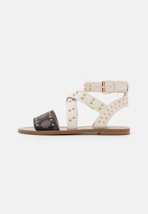 Sandalia de cuero blanca con puntera negra estampada. Presenta tachuelas doradas y un diseño de tiras cruzadas con hebilla ajustable. Suela plana.