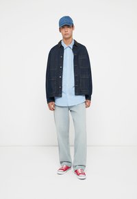 Denim jacket with pockets, light blue striped shirt, light blue jeans, and red sneakers, completed with a blue cap.