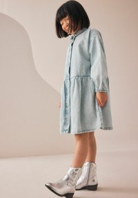 Abito in denim azzurro chiaro con bottoni sul davanti, maniche lunghe e vita arricciata. Abbinato a stivaletti alla caviglia in argento lucido. Texture liscia.