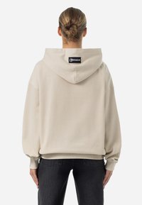 Felpa oversize beige con cappuccio regolabile, polsini e orli a costine. Presenta un'etichetta con logo nera sul retro. Tessuto a trama liscia.