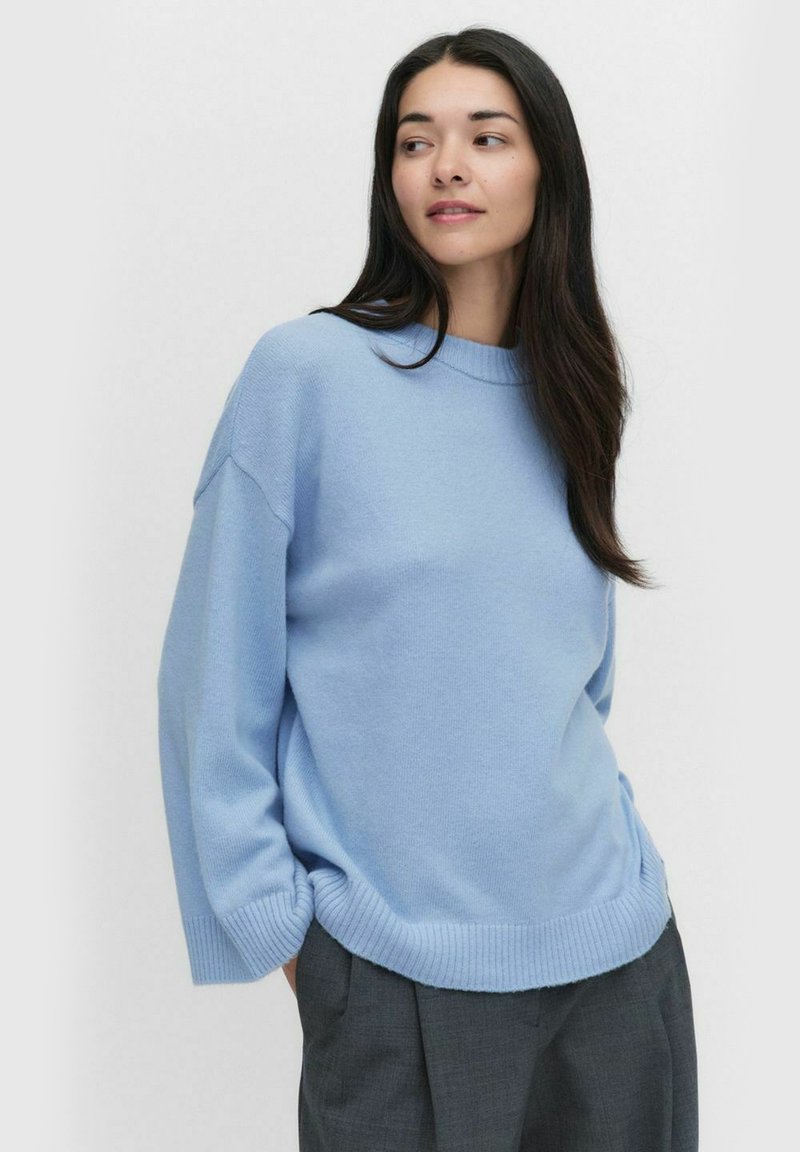 Jasnoniebieski oversize sweter z prążkowanymi mankietami i dołem, miękka tekstura, okrągły dekolt oraz luźny krój, zestawiony z ciemnoszarymi spodniami.