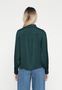 Mørkegrønn satengbluse med krage, lange ermer og knappebufter; har en yoke-detalj og en litt avslappet passform.