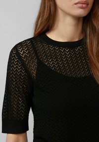 Zwarte gebreide pullover met een zigzagpatroon, met korte mouwen en een ronde hals. De stof is textuur en doorschijnend.
