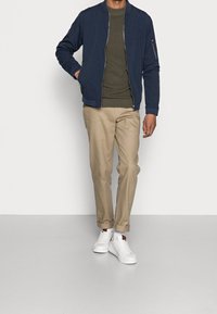 Marineblauer Bomberjacke über einem olivgrünen Hemd, kombiniert mit hochgekrempelten beigefarbenen Chinos und weißen Turnschuhen. Glatter Stoff, Reißverschlusstaschen-Detail.