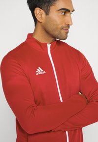 Czerwona kurtka sportowa z wysokim kołnierzem, białym zamkiem i subtelną teksturą. Posiada logo Adidas na górnej lewej części klatki piersiowej.