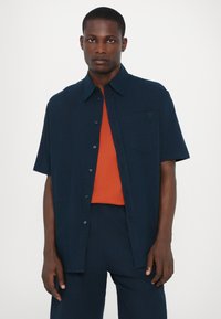Chemise à manches courtes de couleur marine avec un design à boutons, dotée d'une poche poitrine et d'une marque discrète, portée sur une chemise orange.