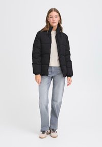 Svart pufferjakke med høy krage og ribbede mansjetter, parret med en beige strikkegenser og lyseblå jeans med rett passform.