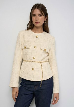 Mujer con chaqueta de tweed color crema con botones dorados y cinturón de cadena, combinada con vaqueros azul oscuro, posando frente a un fondo claro y sencillo.