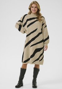Vrouw in een beige midi trui-jurk met zwarte abstracte strepen en zwarte kniehoge laarzen, staande met één hand op de heup.