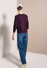 Chemise à manches longues bordeaux, jeans bleu décontractés, et baskets beiges, vue de dos sur un fond neutre.