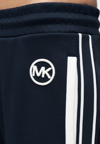 Σκούρο μπλε αθλητικό παντελόνι με λευκές ρίγες στο πλάι και κεντημένο κυκλικό λογότυπο MK. Ομαλό ύφασμα με εφαρμοστή μέση και υφασμάτινες λεπτομέρειες.