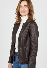 Frau mit schulterlangen, hellbraunen Haaren, die eine dunkelbraune Lederjacke, einen weißen Rollkragenpullover und blaue Jeans trägt und sanft lächelt.