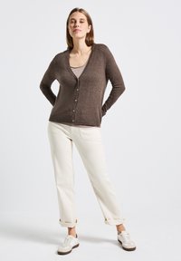 Brun stickad cardigan med V-ringning och framknäppning, kombinerad med ljusa byxor och sneakers. Slät textur.