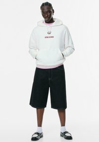 Ung mand med fletninger iført en hvid hoodie med teksten "STWD STUDIO", sorte lange shorts, hvide sokker og sorte slip-on sko.