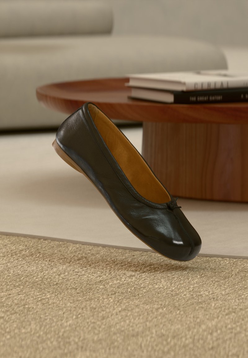 Chaussure ballerine plate en cuir noir avec petit détail nœud, flottant au-dessus d'un tapis beige tissé dans un salon avec une table basse en bois et des livres.
