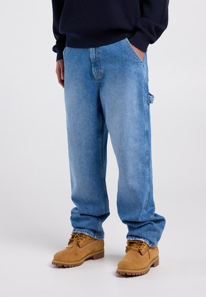 Pantalon en denim bleu clair à coupe ample, avec des poches latérales et une surface texturée. Porté avec des bottines de travail couleur fauve.