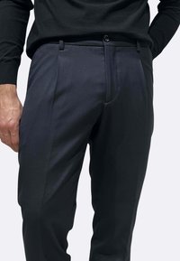 Pantaloni su misura di colore grigio scuro realizzati in tessuto liscio. Presentano una parte anteriore piatta, passanti per cintura e una chiusura con un solo bottone in vita.