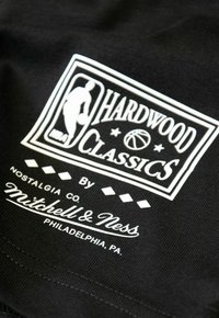 Zwarte stof met een wit logo waarop "HARDWOOD CLASSICS" staat, vergezeld van een basketbalfoto, en de tekst "door Mitchell & Ness, Philadelphia, PA."