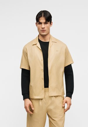 Homme portant une chemise à manches courtes beige à fines rayures et un pantalon assorti par-dessus une chemise noire à manches longues, debout devant un fond uni.