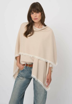 Frau mit langem braunem Haar, die einen beigefarbenen Poncho mit Fransen über blauen Jeans und einem braunen Gürtel trägt und vor einem einfarbigen Hintergrund steht.