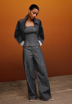 Vrouw draagt donkere denim broek met wijde pijpen, strapless top en bijpassend jasje met omgeslagen manchetten, poserend tegen een bruine achtergrond.